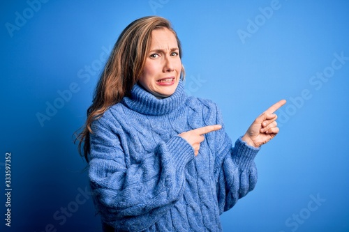 Young beautiful blonde woman wearing casual turtleneck sweater over blue background Pointing aside worried and nervous with both hands, concerned and surprised expression