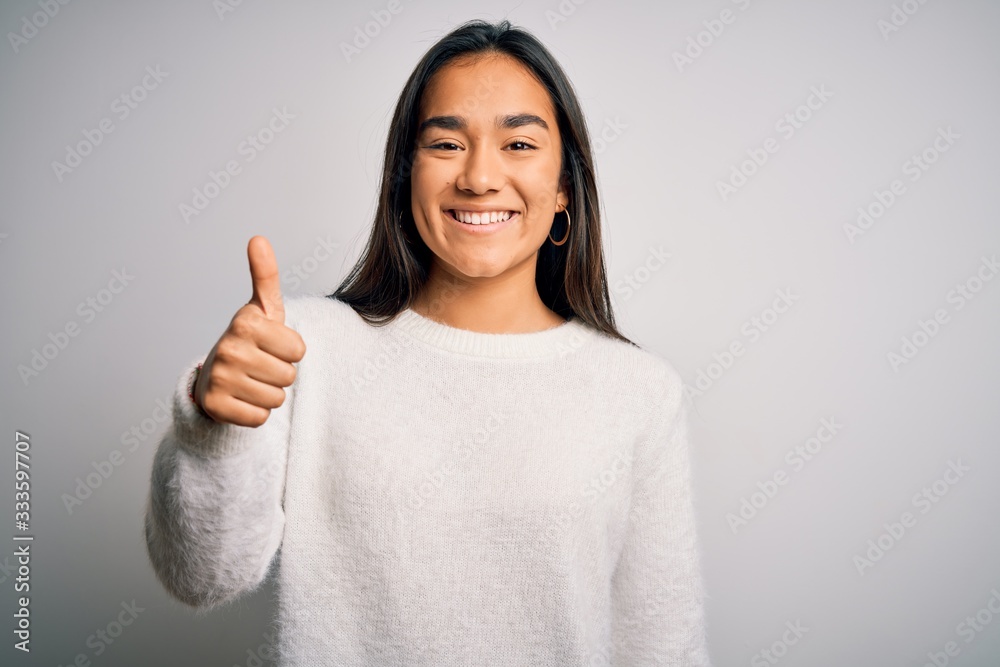 Young beautiful asian woman wearing casual sweater standing over white background doing happy thumbs up gesture with hand. Approving expression looking at the camera showing success. Young beautiful asian woman wearing casual sweater standing over white background doing happy thumbs up gesture with hand. Approving expression looking at the camera showing success.
