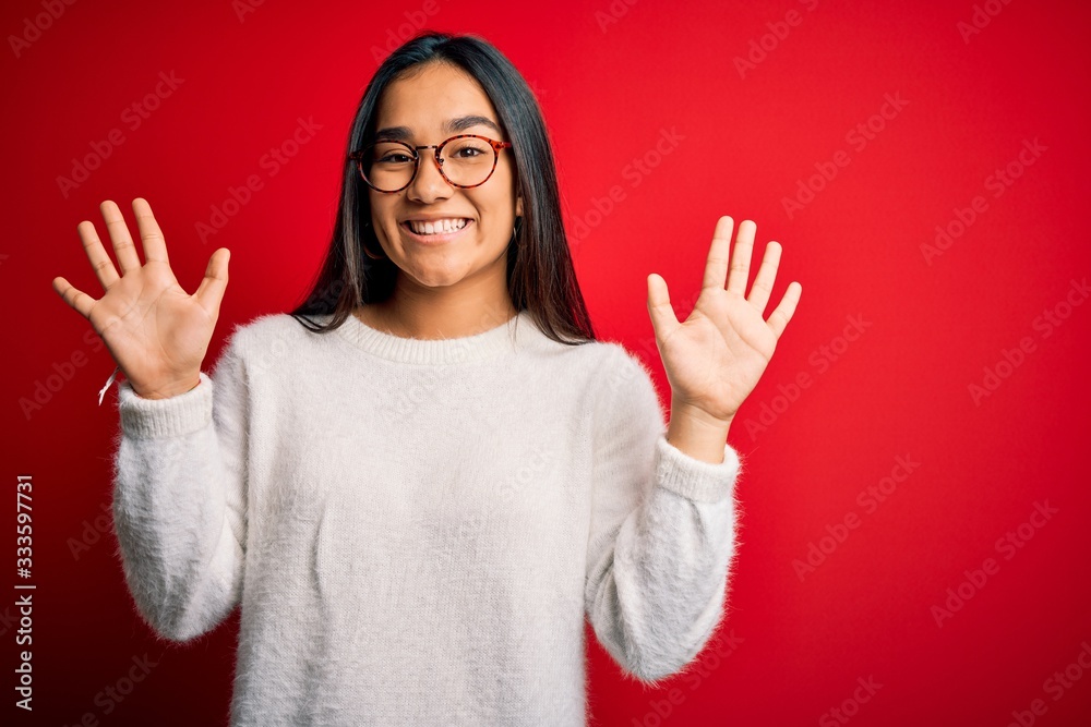 Young beautiful asian woman wearing casual sweater and glasses over red background showing and pointing up with fingers number ten while smiling confident and happy. Young beautiful asian woman wearing casual sweater and glasses over red background showing and pointing up with fingers number ten while smiling confident and happy.
