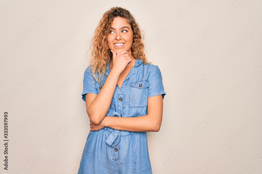 Young beautiful woman with blue eyes wearing casual denim dress over white background with hand on chin thinking about question, pensive expression. Smiling with thoughtful face. Doubt concept. Young beautiful woman with blue eyes wearing casual denim dress over white background with hand on chin thinking about question, pensive expression. Smiling with thoughtful face. Doubt concept.