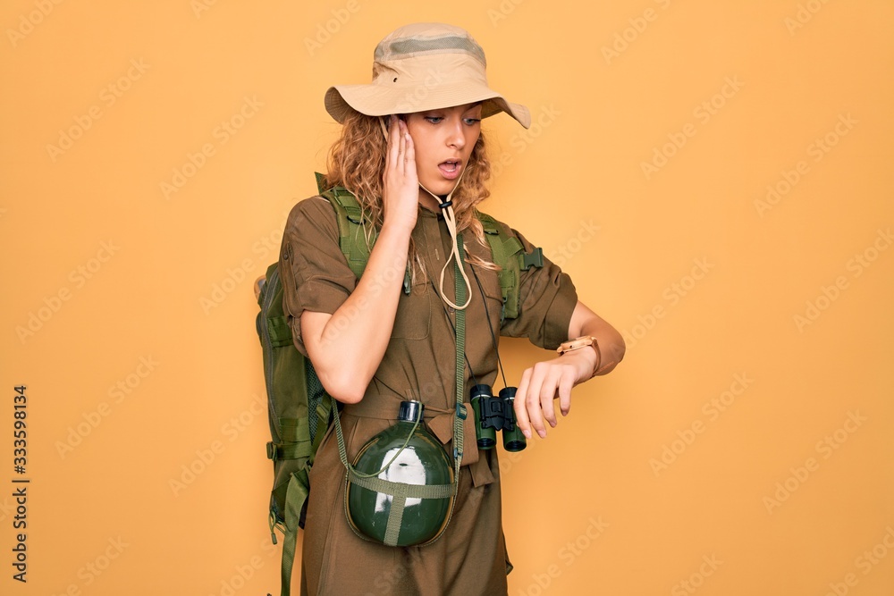 Young blonde explorer woman with blue eyes hiking wearing backpack and water canteen Looking at the watch time worried, afraid of getting late