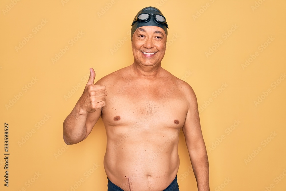 Middle age senior grey-haired swimmer man wearing swimsuit, cap and goggles doing happy thumbs up gesture with hand. Approving expression looking at the camera showing success.