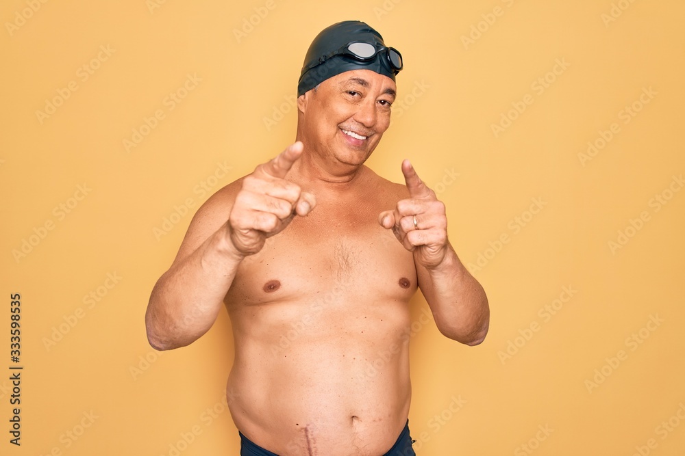 Middle age senior grey-haired swimmer man wearing swimsuit, cap and goggles pointing fingers to camera with happy and funny face. Good energy and vibes.