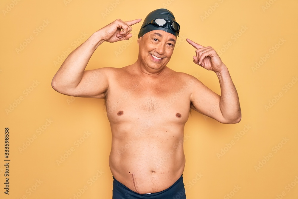 Middle age senior grey-haired swimmer man wearing swimsuit, cap and goggles smiling pointing to head with both hands finger, great idea or thought, good memory