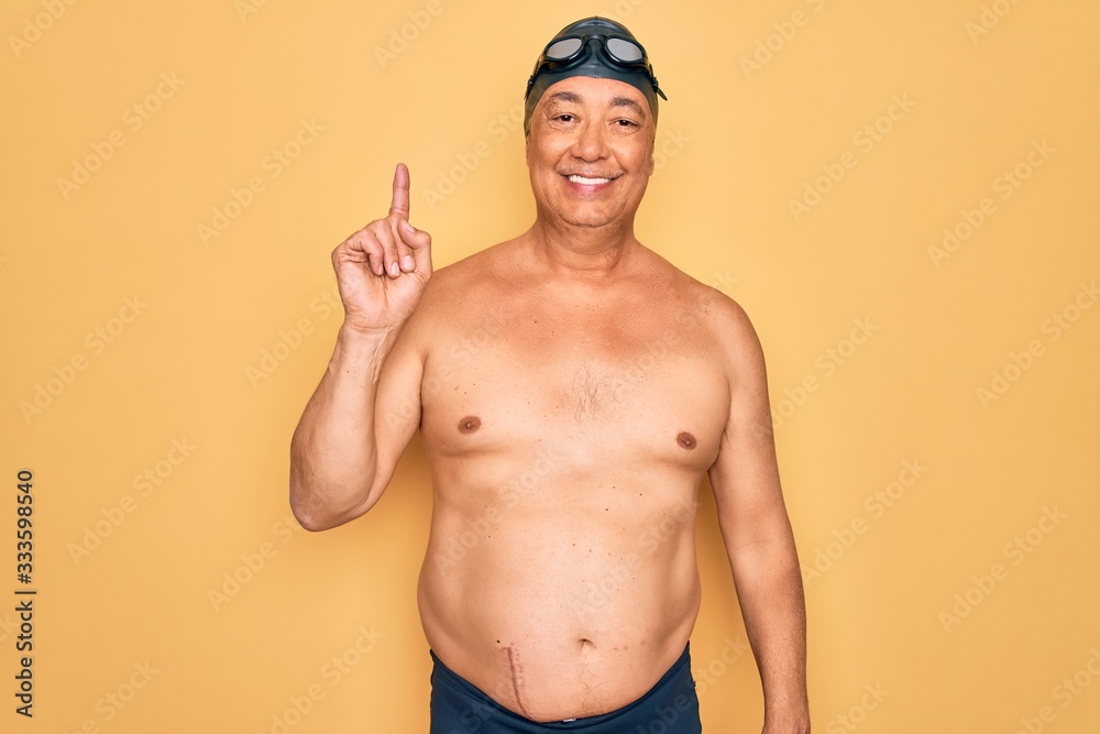 Middle age senior grey-haired swimmer man wearing swimsuit, cap and goggles showing and pointing up with finger number one while smiling confident and happy.
