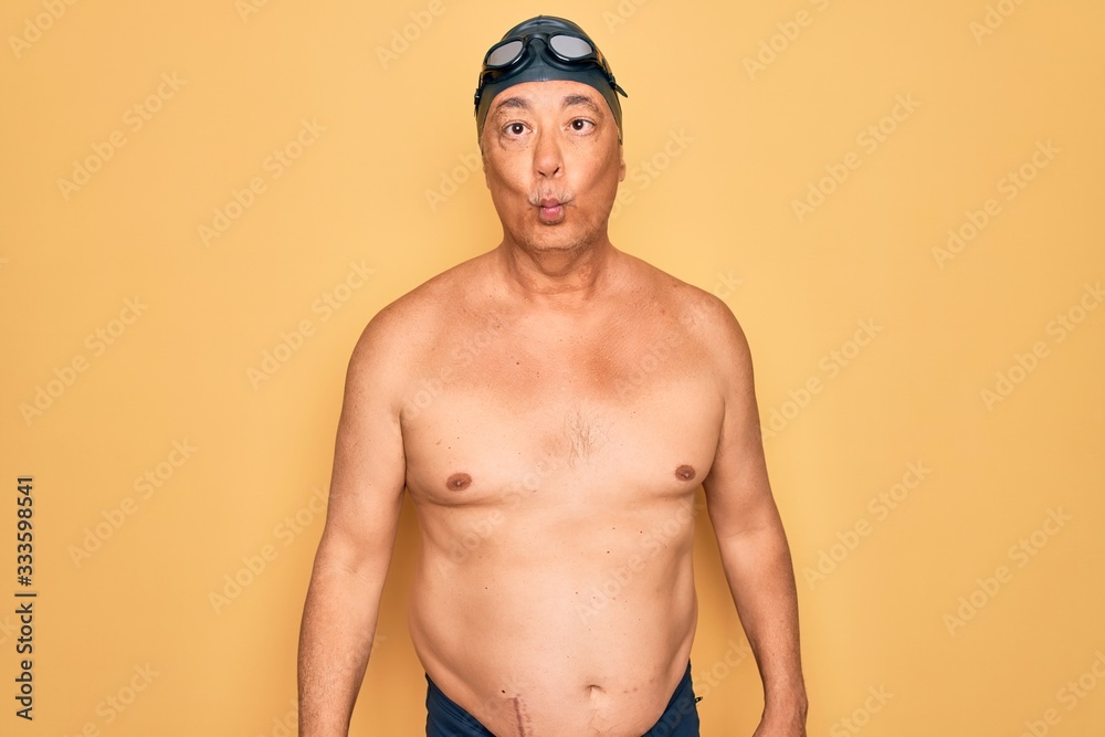 Middle age senior grey-haired swimmer man wearing swimsuit, cap and goggles making fish face with lips, crazy and comical gesture. Funny expression.