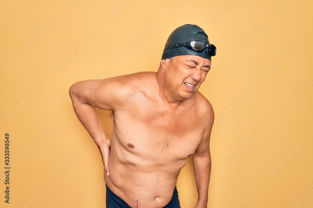 Middle age senior grey-haired swimmer man wearing swimsuit, cap and goggles Suffering of backache, touching back with hand, muscular pain