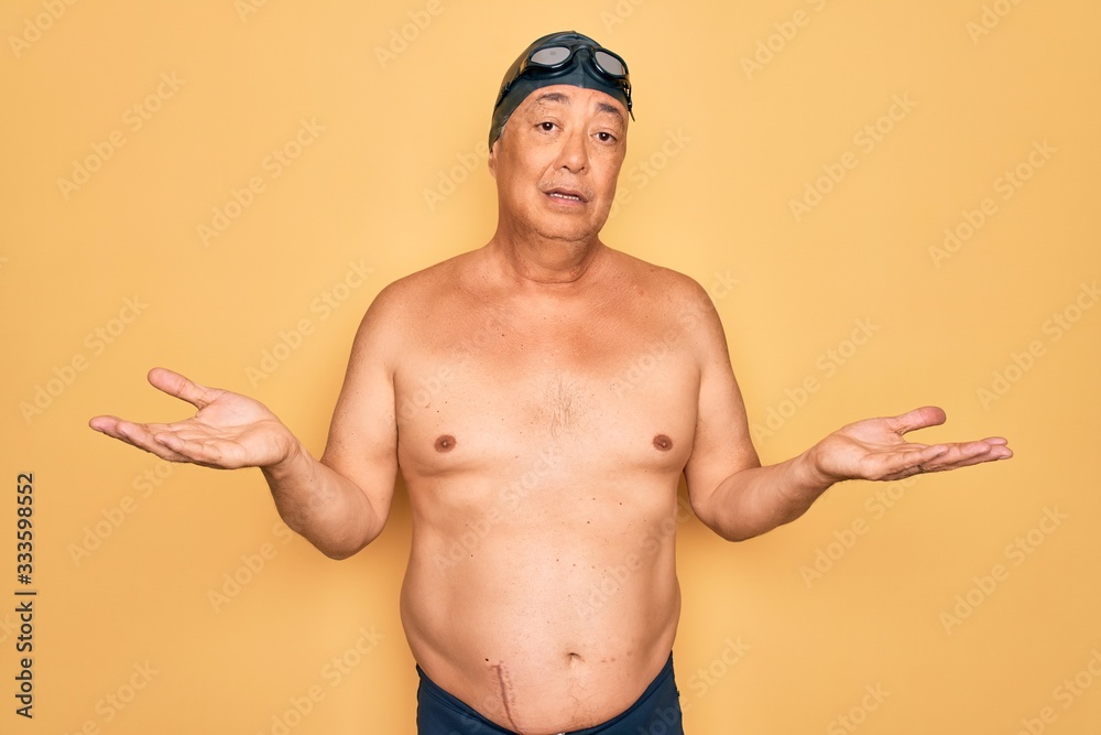 Middle age senior grey-haired swimmer man wearing swimsuit, cap and goggles clueless and confused with open arms, no idea concept.
