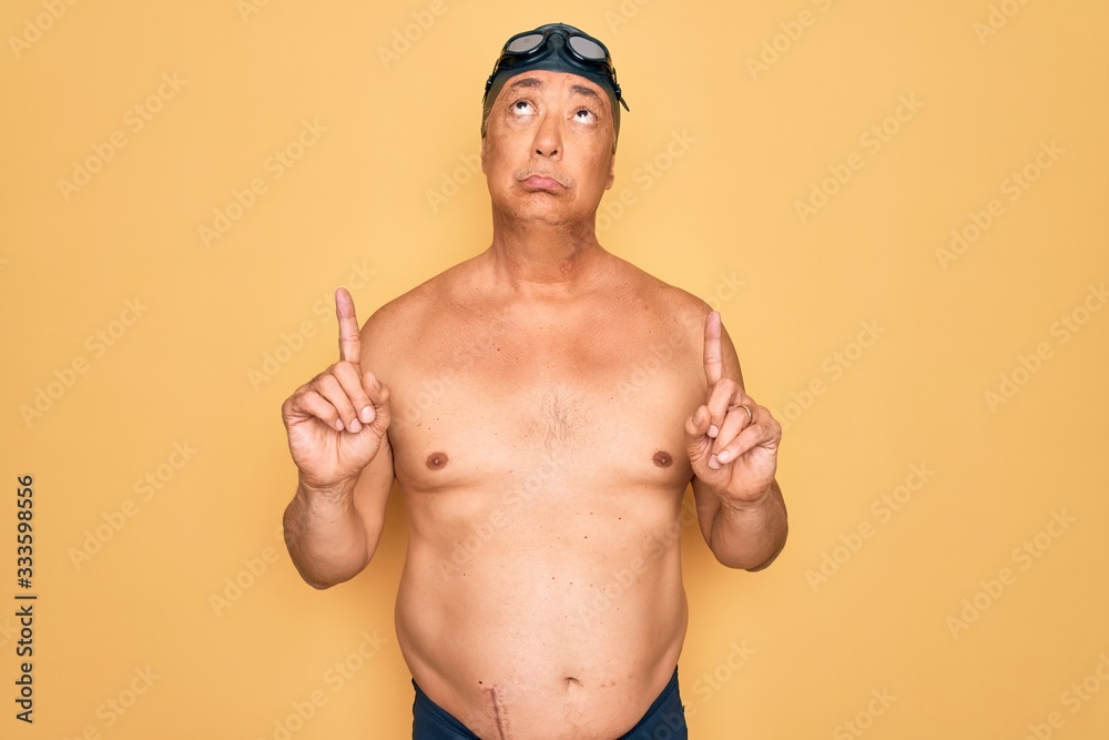 Middle age senior grey-haired swimmer man wearing swimsuit, cap and goggles Pointing up looking sad and upset, indicating direction with fingers, unhappy and depressed.