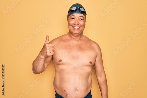 Middle age senior grey-haired swimmer man wearing swimsuit  cap and goggles doing happy thumbs up gesture with hand. Approving expression looking at the camera showing success.