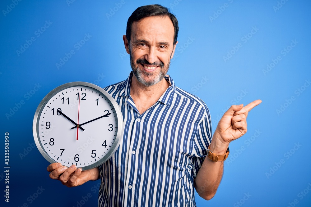 Middle age hoary man holding big clock standing over isolated blue background very happy pointing with hand and finger to the side Middle age hoary man holding big clock standing over isolated blue background very happy pointing with hand and finger to the side