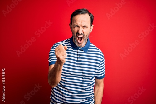 Middle age hoary man wearing casual striped polo standing over isolated red background angry and mad raising fist frustrated and furious while shouting with anger. Rage and aggressive concept.