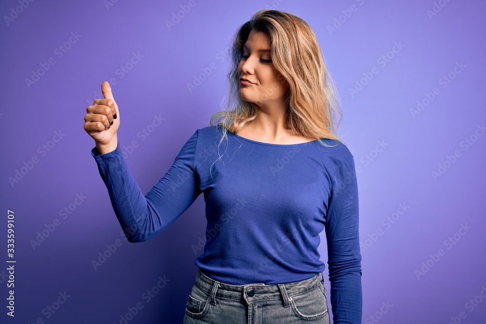Young beautiful blonde woman wearing casual t-shirt over isolated purple background Looking proud, smiling doing thumbs up gesture to the side Young beautiful blonde woman wearing casual t-shirt over isolated purple background Looking proud, smiling doing thumbs up gesture to the side