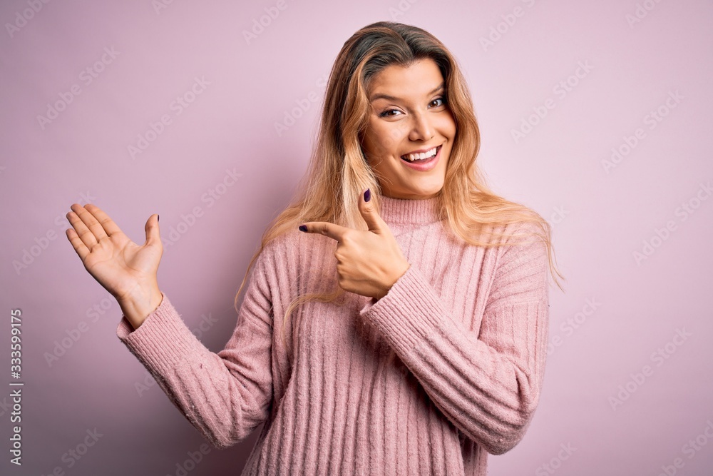 Young beautiful blonde woman wearing casual pink sweater over isolated background amazed and smiling to the camera while presenting with hand and pointing with finger. Young beautiful blonde woman wearing casual pink sweater over isolated background amazed and smiling to the camera while presenting with hand and pointing with finger.