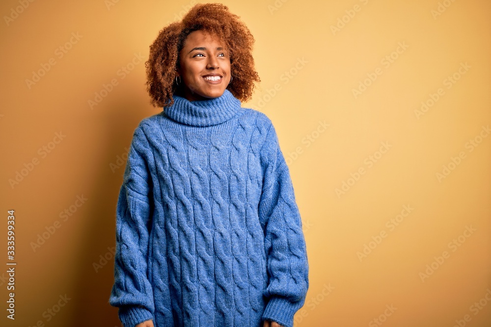 Young beautiful African American afro woman with curly hair wearing blue turtleneck sweater looking away to side with smile on face, natural expression. Laughing confident. Young beautiful African American afro woman with curly hair wearing blue turtleneck sweater looking away to side with smile on face, natural expression. Laughing confident.