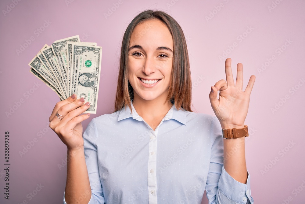 Young beautiful woman holding bunch of dollars banknotes over isolated pink background doing ok sign with fingers, excellent symbol Young beautiful woman holding bunch of dollars banknotes over isolated pink background doing ok sign with fingers, excellent symbol