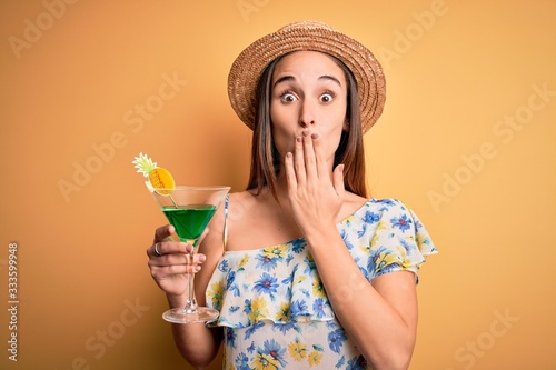 Young beautiful tourist woman on vacation wearing summer hat drinking cocktail beverage cover mouth with hand shocked with shame for mistake, expression of fear, scared in silence, secret concept