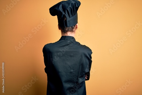 Young beautiful chef woman wearing cooker uniform and hat standing over yellow background standing backwards looking away with crossed arms