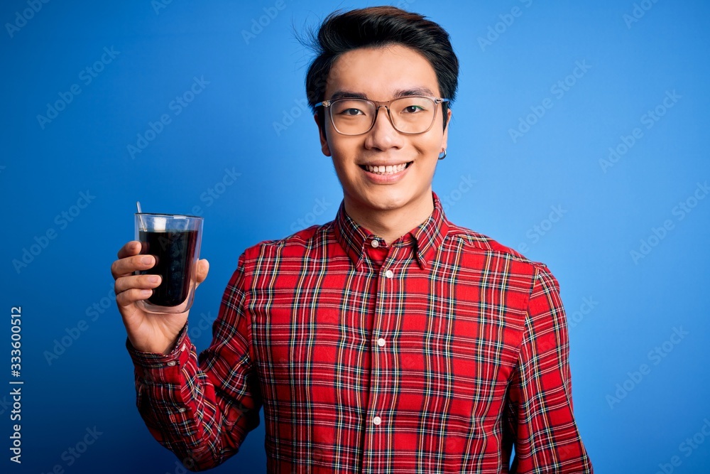 Young handsome chinese man drinking glass of coffee over isolated blue background with a happy face standing and smiling with a confident smile showing teeth Young handsome chinese man drinking glass of coffee over isolated blue background with a happy face standing and smiling with a confident smile showing teeth