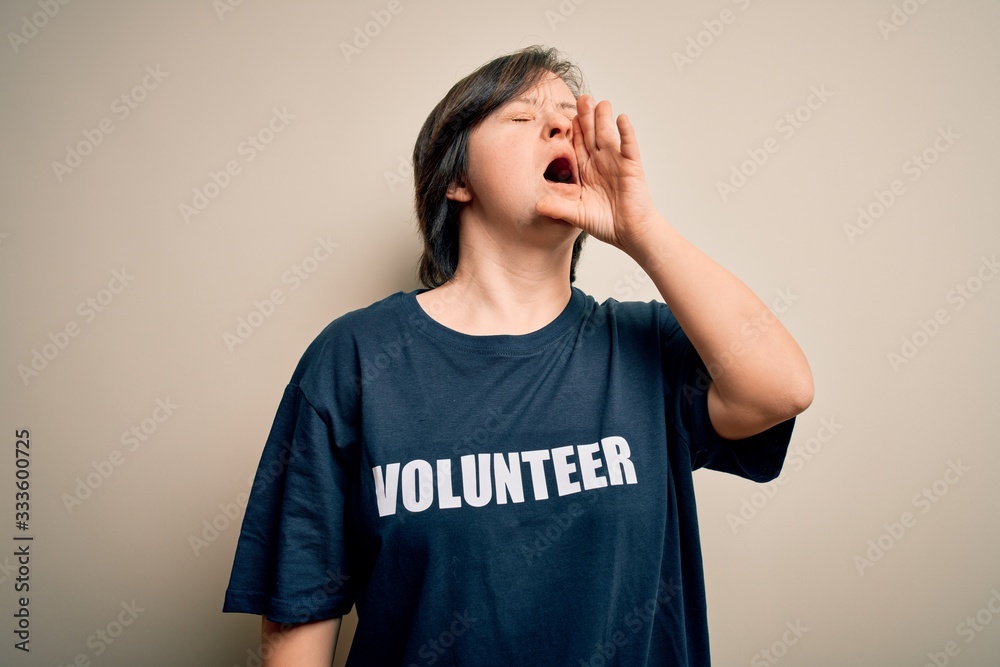 Young down syndrome volunteer woman wearing social care charity t-shirt shouting and screaming loud to side with hand on mouth. Communication concept. Young down syndrome volunteer woman wearing social care charity t-shirt shouting and screaming loud to side with hand on mouth. Communication concept.
