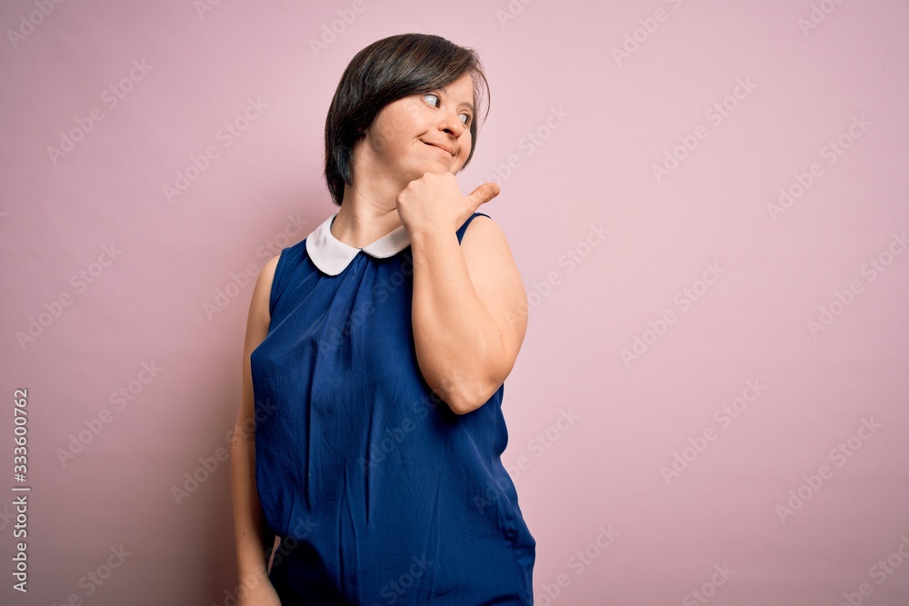 Young down syndrome woman wearing elegant shirt over pink background smiling with happy face looking and pointing to the side with thumb up. Young down syndrome woman wearing elegant shirt over pink background smiling with happy face looking and pointing to the side with thumb up.