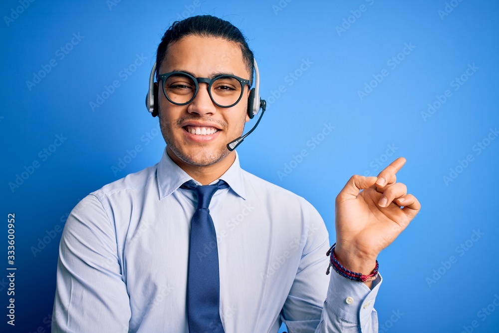 Young brazilian call center agent man wearing glasses and tie working using headset with a big smile on face, pointing with hand and finger to the side looking at the camera. Young brazilian call center agent man wearing glasses and tie working using headset with a big smile on face, pointing with hand and finger to the side looking at the camera.