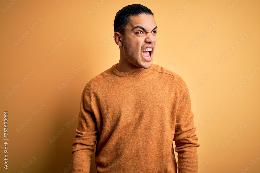 Young brazilian man wearing casual sweater standing over isolated yellow background angry and mad screaming frustrated and furious, shouting with anger. Rage and aggressive concept. Young brazilian man wearing casual sweater standing over isolated yellow background angry and mad screaming frustrated and furious, shouting with anger. Rage and aggressive concept.