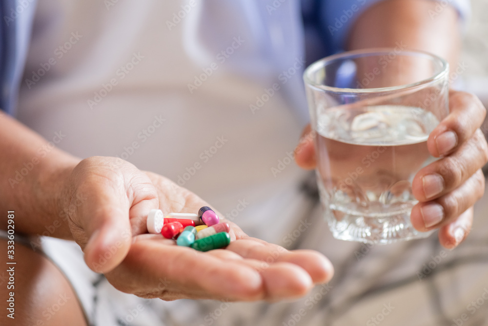 Asian old man taking in pill and another hand holding a glass of clean mineral water. Senior healthcare and medicine concept.