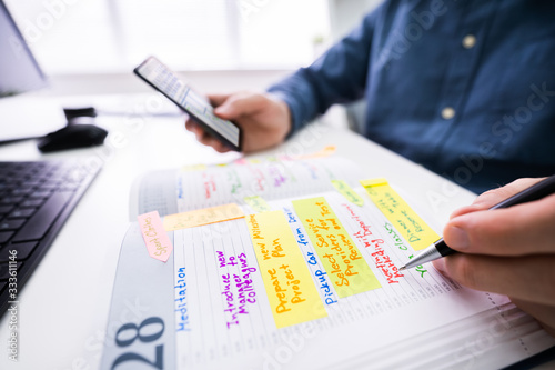 Businessman Holding Cellphone Writing Schedule In Diary With Pen