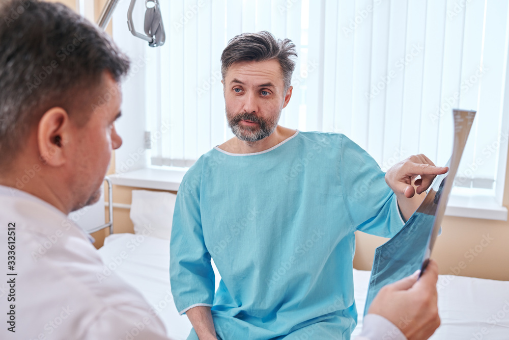 Mature bearded patient in hospital gown sitting on bed and pointing at x-ray image while asking doctor about his disease Mature bearded patient in hospital gown sitting on bed and pointing at x-ray image while asking doctor about his disease