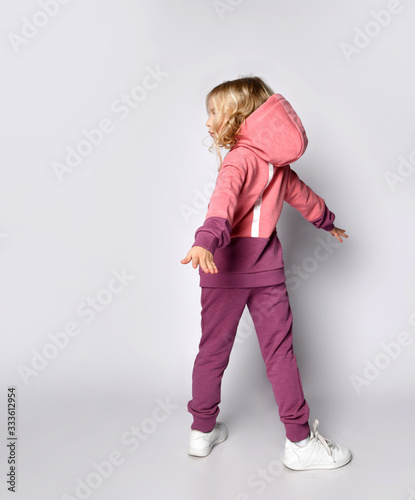 Curly hair blonde kid girl in modern fashion pink gray sportsuit stands back to us with her arms outstretched spinning