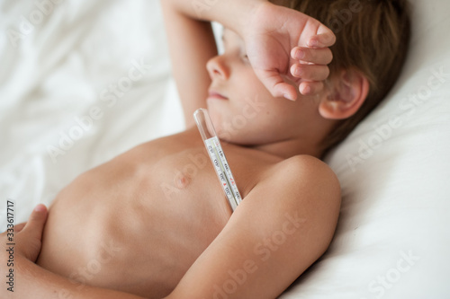 healthcare quarantine concept of sick little boy with high temperature lying in bed with thermometer under arm during covid-19 pandemic outbreak epidemic spead