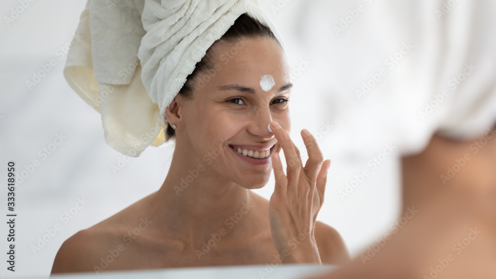 Woman reflected in mirror after morning shower with towel on head applies day facial cream close up, prevent first wrinkles, caring about skin use moisturizer creme, beauty treatment, skincare concept Woman reflected in mirror after morning shower with towel on head applies day facial cream close up, prevent first wrinkles, caring about skin use moisturizer creme, beauty treatment, skincare concept