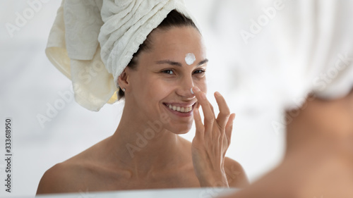 Woman reflected in mirror after morning shower with towel on head applies day facial cream close up, prevent first wrinkles, caring about skin use moisturizer creme, beauty treatment, skincare concept