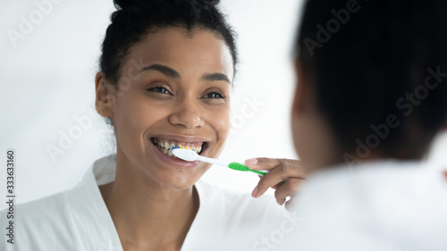 Personal oral hygiene and dental care concept. African ethnicity beautiful 30s woman woke up in morning looks in mirror holds toothbrush brushing her teeth smiles enjoy morning routine regular process