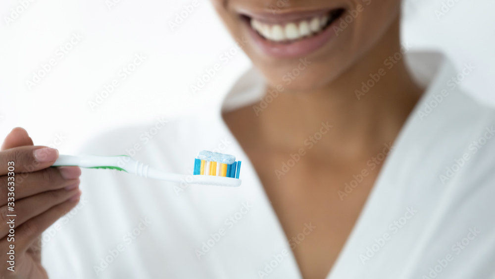 Close up African woman holding toothbrush with toothpaste, on background charming perfect smile of American female. Everyday routine, self-care, teeth cleaning, oral hygiene caries prevention concept Close up African woman holding toothbrush with toothpaste, on background charming perfect smile of American female. Everyday routine, self-care, teeth cleaning, oral hygiene caries prevention concept