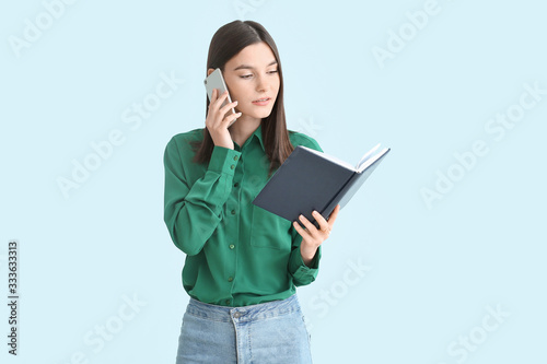 Beautiful young woman with book talking by mobile phone on color background