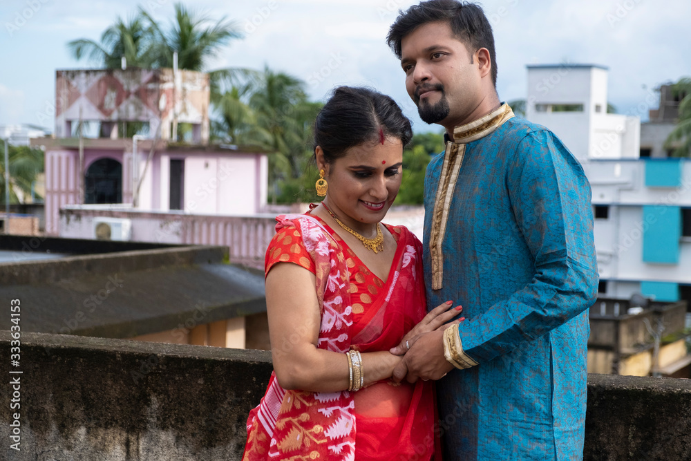 Naklejka premium A brunette Indian Bengali romantic couple in traditional wear enjoying themselves standing on the roof top in sunny morning of Durga Puja festival in green natural background. Indian lifestyle.