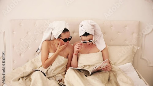 Portrait of thwo friends with white towels on their heads and with sunglasses lie on a bed under the white blanket, read magazines and surprised by something photo