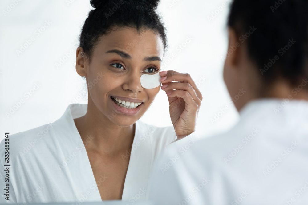 Awakened African woman in white bathrobe looking in mirror solves under eyes facial problems applies patches to reduce dark circles, remove fine wrinkles. Beauty treatment, skin care, grooming concept Awakened African woman in white bathrobe looking in mirror solves under eyes facial problems applies patches to reduce dark circles, remove fine wrinkles. Beauty treatment, skin care, grooming concept