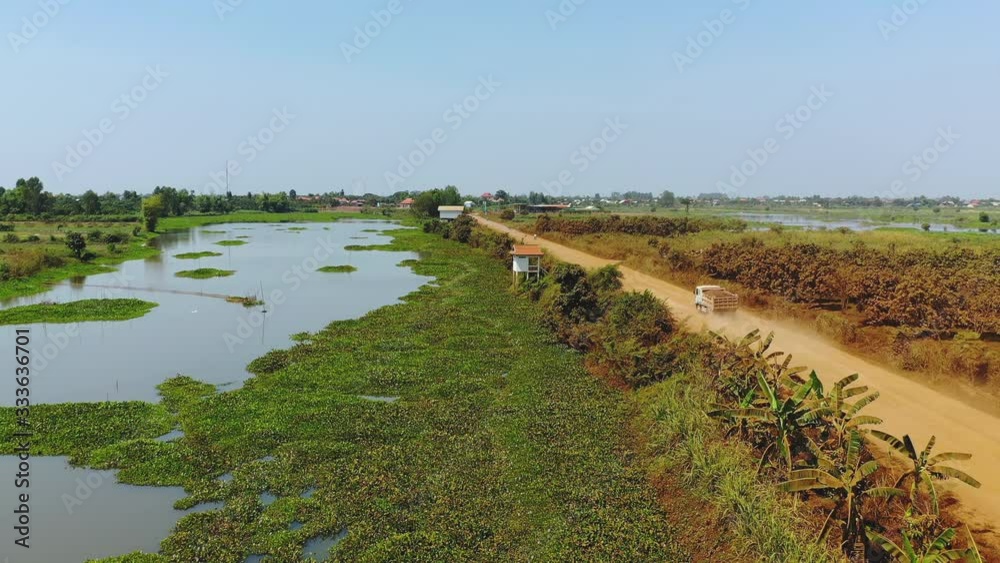 Un camion et quelques motos circulent sur une route de terre et de sable orange au bord d'un petit étang en faisant beaucoup de poussière vers Phnom Penh, au Cambodge, en Asie.