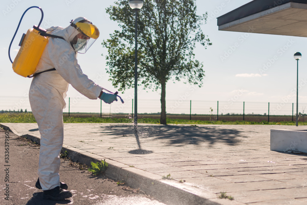 Man wearing an NBC personal protective equipment (ppe) suit, gloves, mask, and face shield, cleaning the streets with a backpack of pressurized spray disinfectant water to remove covid-19 coronavirus. Man wearing an NBC personal protective equipment (ppe) suit, gloves, mask, and face shield, cleaning the streets with a backpack of pressurized spray disinfectant water to remove covid-19 coronavirus.