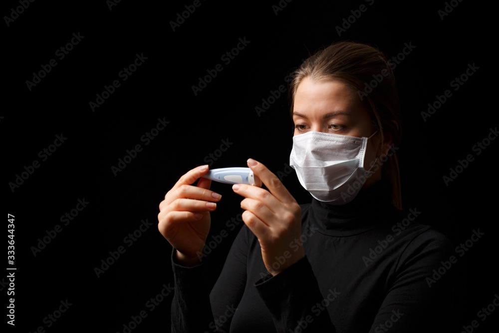 Coronavirus woman with a mask in looks at a thermometer. Studio portrait on a black background.
