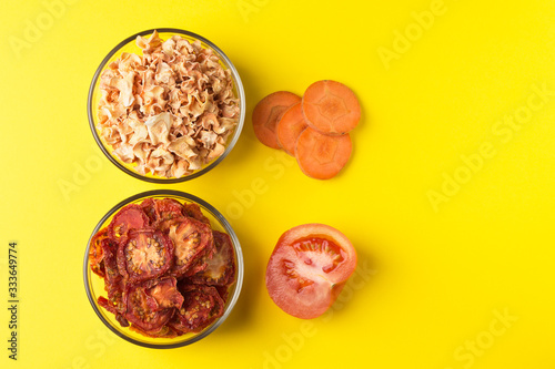 Dried tomato and carrot slices in a glass dish on a bright yellow background. There are fresh pieces of vegetables nearby.