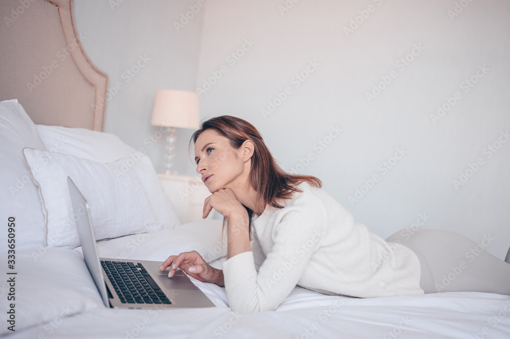 Young european woman working on a laptop in white bedroom during coronavirus isolation home quarantine. Pandemic Corona virus. Distance online work from home. COVID-19 concept promote stay safe home Young european woman working on a laptop in white bedroom during coronavirus isolation home quarantine. Pandemic Corona virus. Distance online work from home. COVID-19 concept promote stay safe home