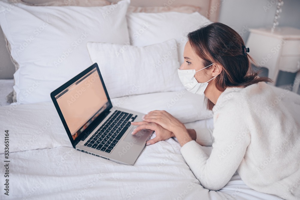 Young european woman in face medicine mask working on a laptop in bedroom during coronavirus isolation home quarantine. Covid-19 pandemic Corona virus. Distance online work from home concept.