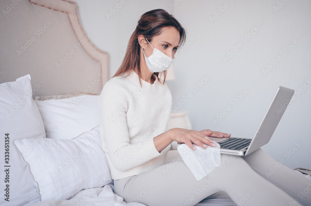 European woman in face mask in bedroom during coronavirus isolation home quarantine cleaning laptop by hand sanitizer, using cotton wool with alcohol to wipe to avoid contaminating with Corona virus. European woman in face mask in bedroom during coronavirus isolation home quarantine cleaning laptop by hand sanitizer, using cotton wool with alcohol to wipe to avoid contaminating with Corona virus.