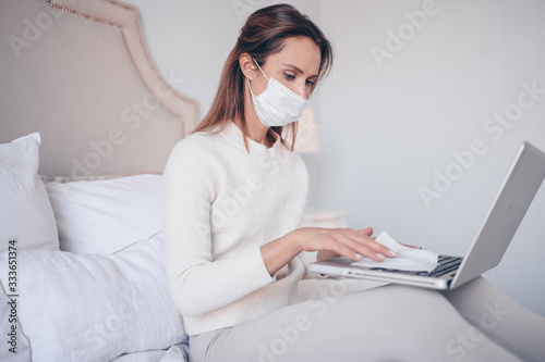 European woman in face mask in bedroom during coronavirus isolation home quarantine cleaning laptop by hand sanitizer, using cotton wool with alcohol to wipe to avoid contaminating with Corona virus.