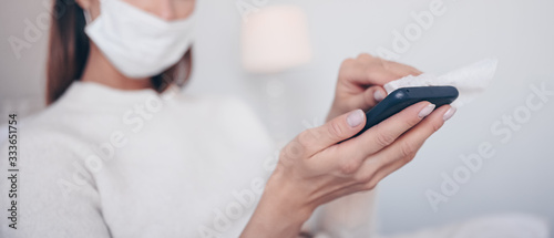 Unrecognizable woman in protection face mask  during coronavirus isolation home quarantine cleaning phone by hand sanitizer, using cotton wool with alcohol. Coronavirus COVID-19 pandemic concept