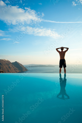 Unrecognizable man standing on the edge of an infinity pool looking out over a dramatic Mediterranean view of the Santorini caldera, Greece